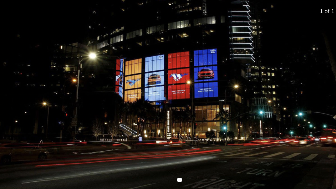 City intersection at night with digital screens on a high-rise building showing Ford Mustang advertisements.