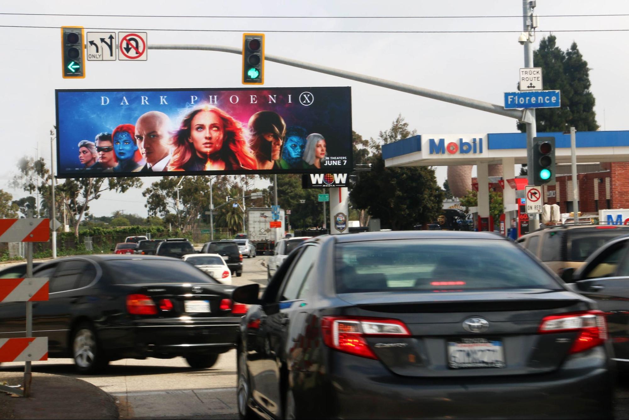 dark-phoenix-billboard Street intersection with a digital billboard displaying the 'Dark Phoenix' movie poster featuring the main cast, located near a Mobil gas station."