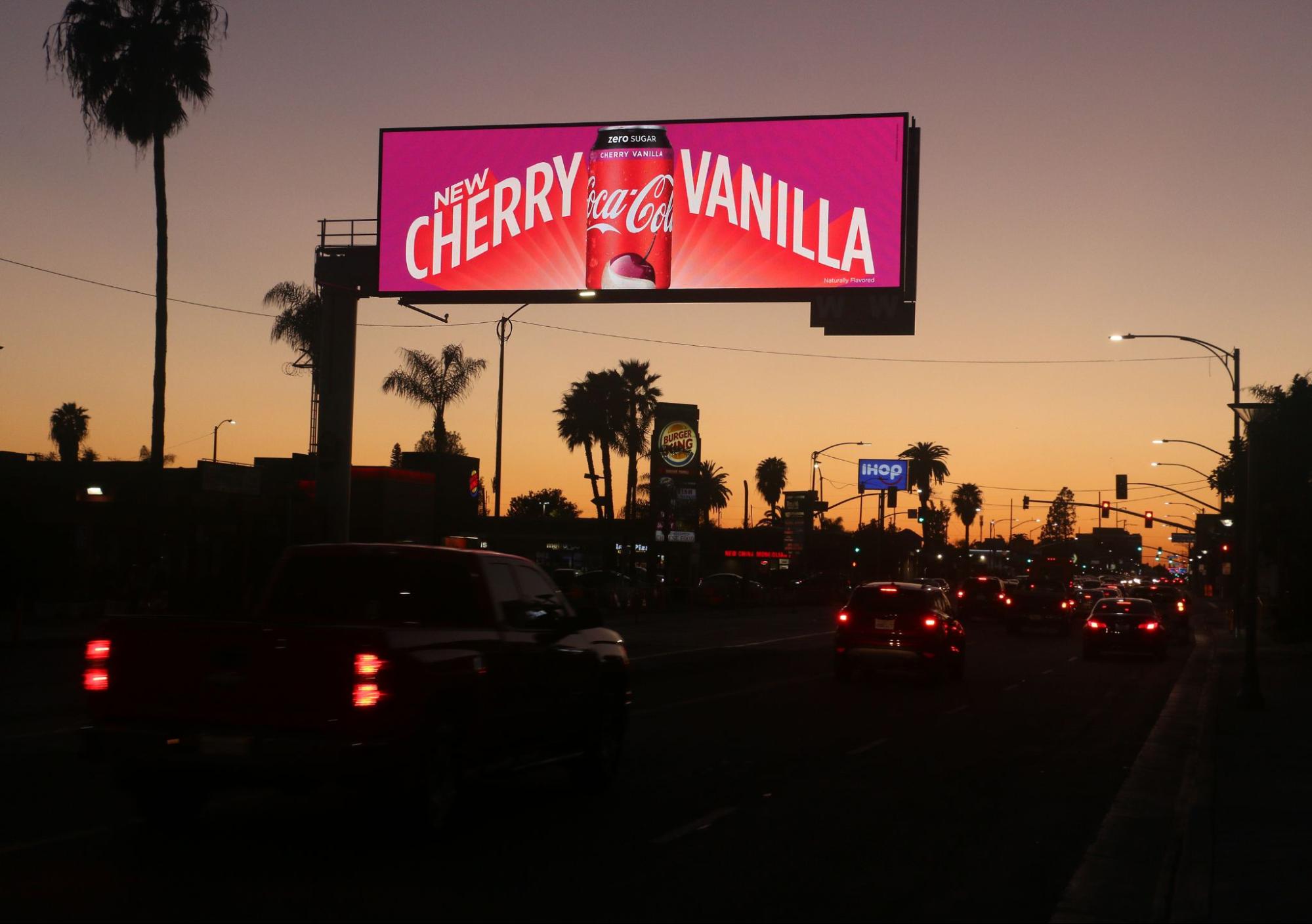 coca-cola-billboard Dusk cityscape with a Coca-Cola Cherry Vanilla digital billboard, cars on the road, and palm trees in the background.