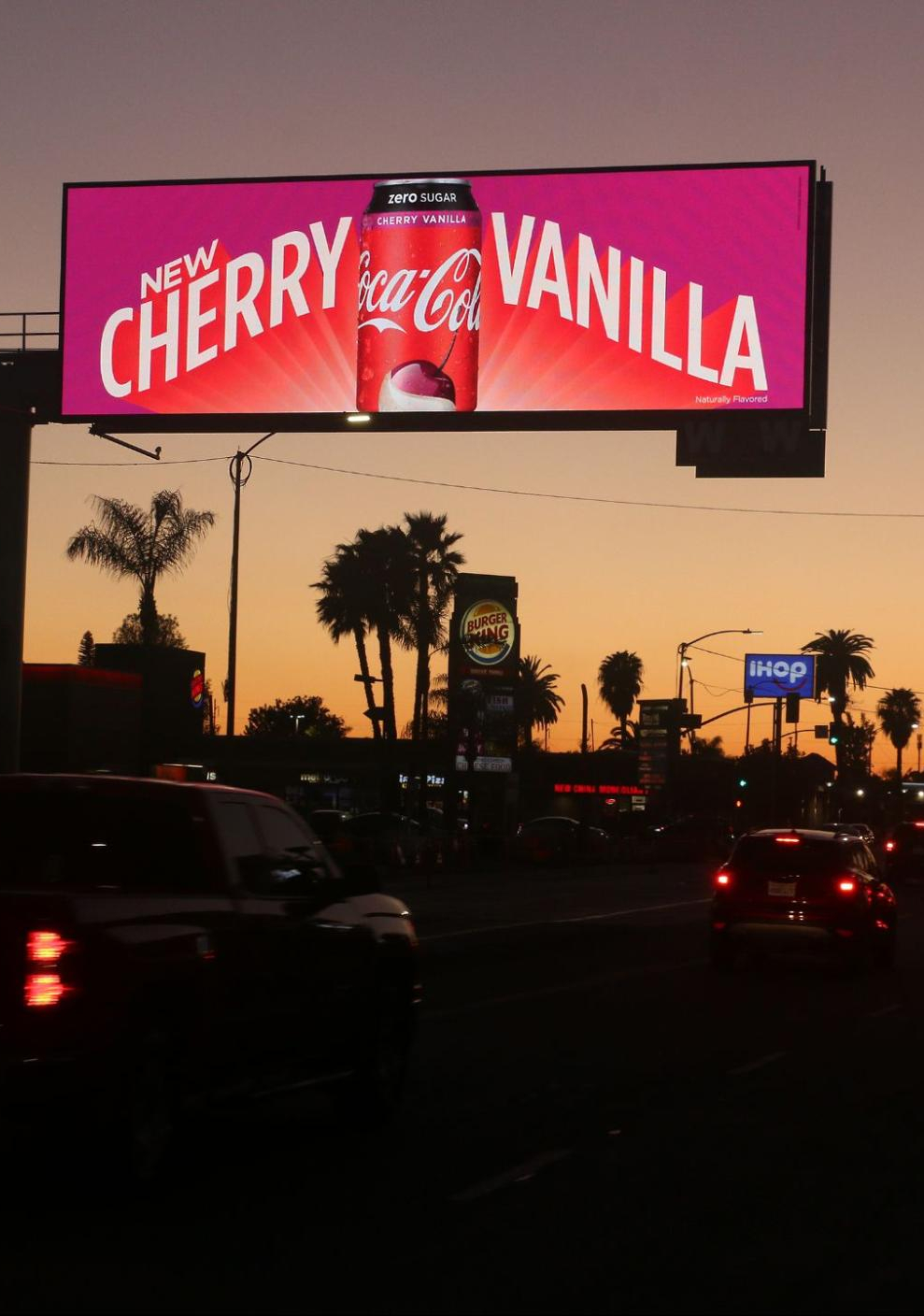 new-cherry-vainilla-billboard City street at dusk with a large Coca-Cola Cherry Vanilla ad displayed on a digital billboard.