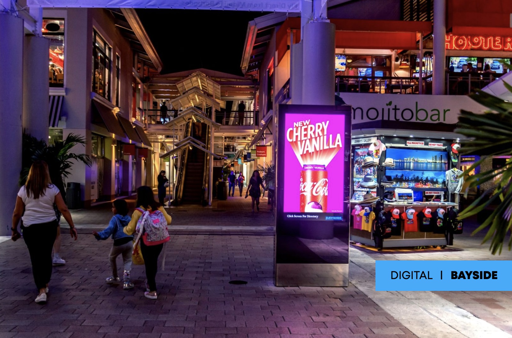 miami-billboard Night scene at Bayside shopping area with a digital billboard displaying Coca-Cola Cherry Vanilla ad, surrounded by shops and visitors.