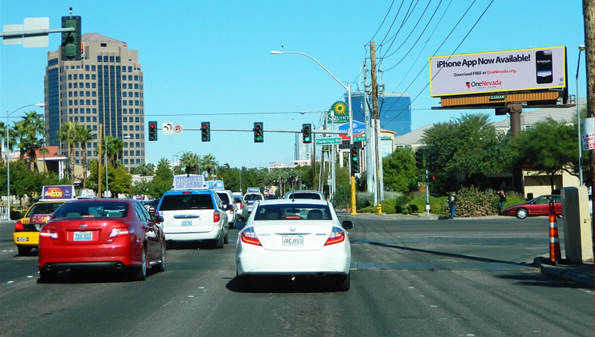 las-vegas-iphone-billboard Traffic at a Las Vegas intersection under a digital billboard advertising One Nevada’s iPhone app, with palm trees and office buildings in the background.